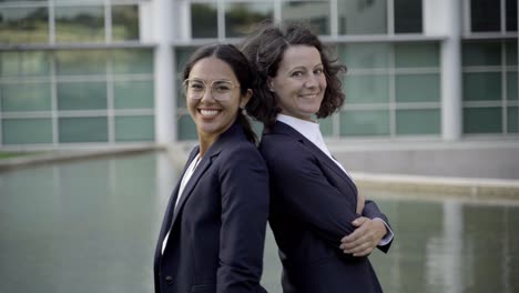 Two-smiling-women-standing-back-to-back-near-office-building