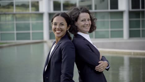 dos mujeres sonrientes de pie de espaldas cerca del edificio de oficinas