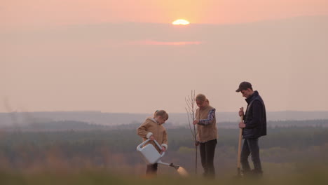 Familie-Mit-Einem-Baumsetzling,-Einem-Sprinkler-Und-Einer-Schaufel,-Die-An-Einem-Malerischen-Ort-Steht