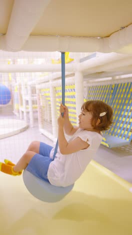 a joyful young girl swings happily on a light blue tire swing on a brightly lit indoor playground, enjoying her playtime