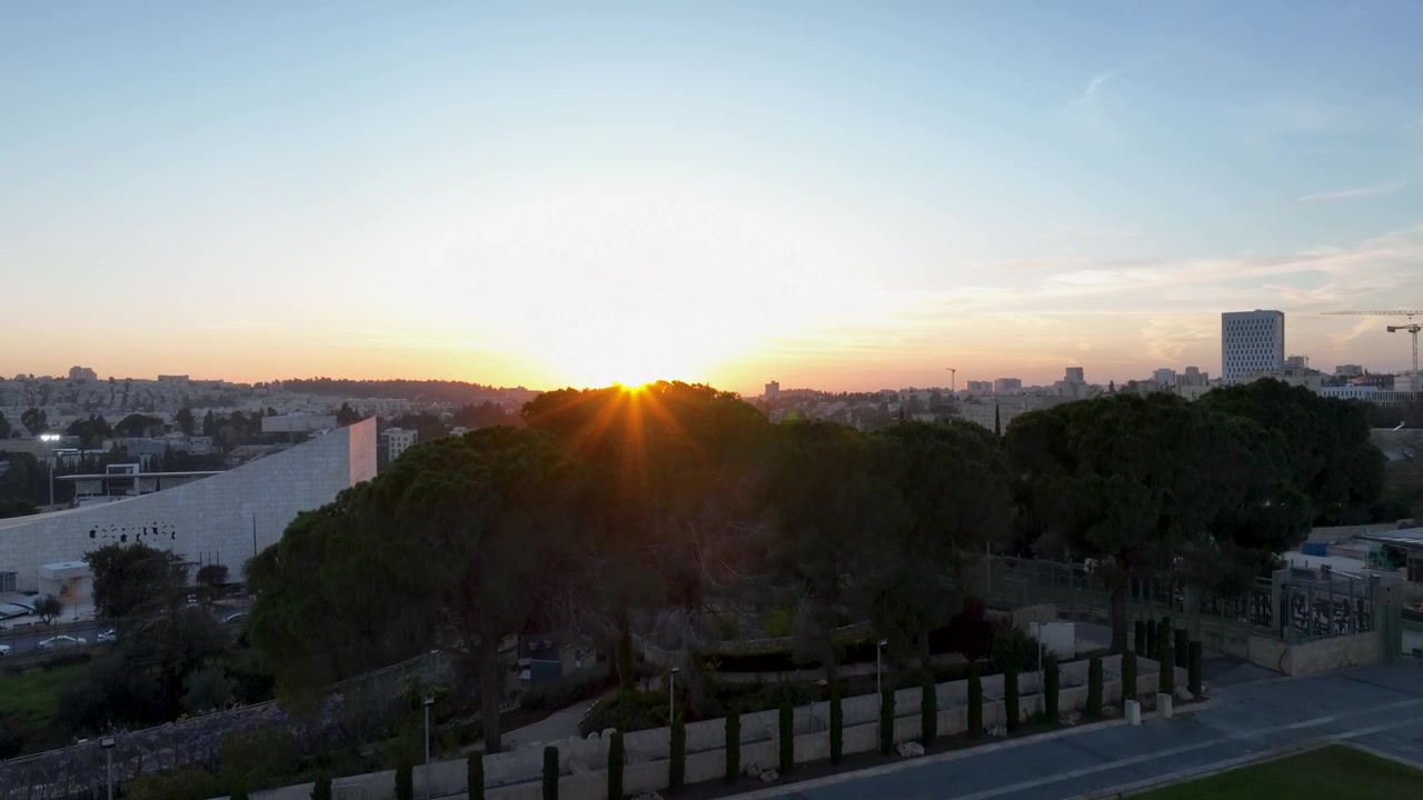Raios quentes do pôr do sol surgindo sobre as copas dos pinheiros e marcos arquitetônicos modernos no horizonte de Jerusalém durante o crepúsculo.