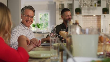 Happy-caucasian-parents,-children-and-grandfather-sitting-at-table-holding-hands-before-family-meal