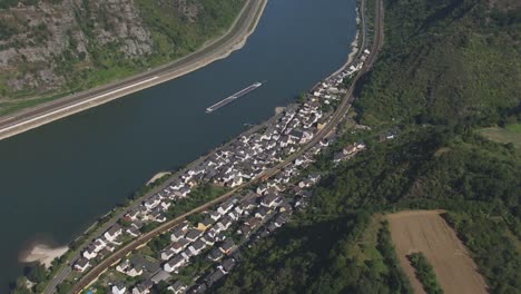 disparo de avión no tripulado capturando una pequeña ciudad con edificios blancos y techos grises enclavado por un río ancho con una barcaza, rodeado de colinas verdes y bosques, dividido en dos por una carretera