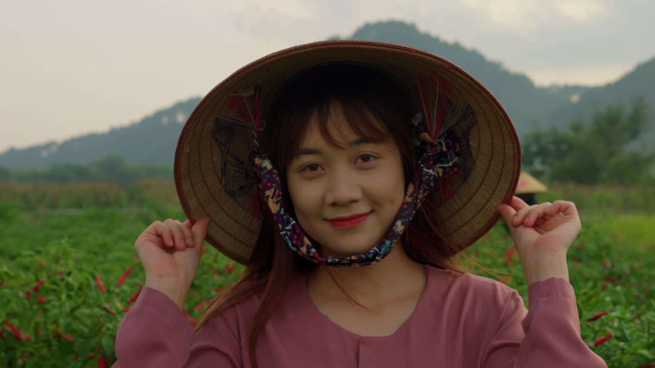 야외에서 일하는 동안 농장 한가운데 서 있는  대나무 모자를 입은 젊은 웃는 중국 농부의 초상화