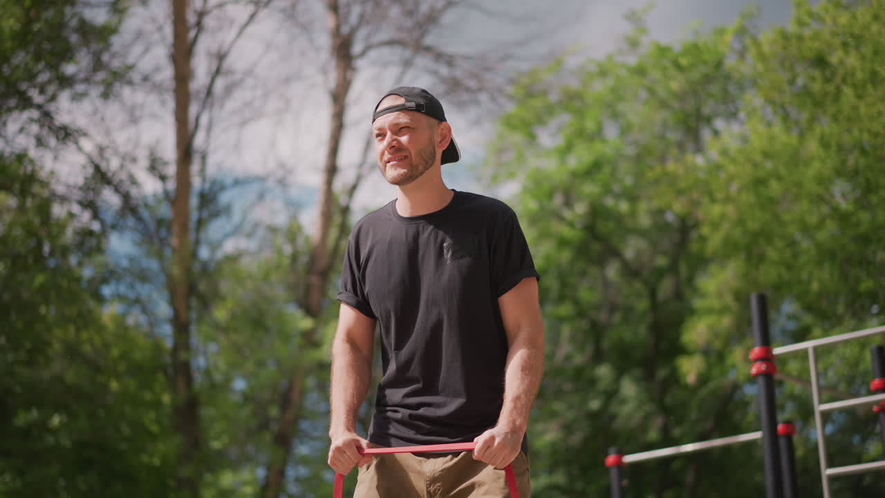 White Male Exercising At Outdoor Gym With Red Band, Trainer Style Demonstration On Bars, Focused Pulls And Controlled Reps, Calisthenics Setting With Metal Bars And Leafy Backdrop, Energetic Summer