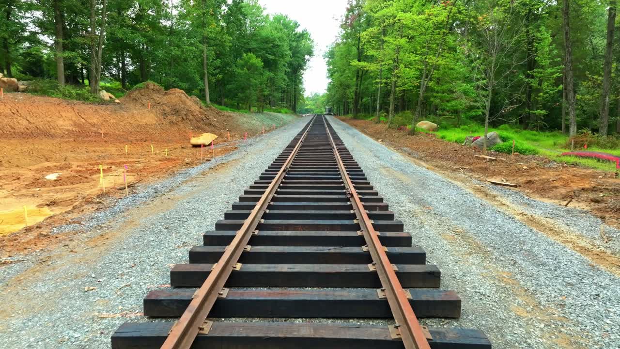 Workers are in the process of laying new railroad tracks in a tranquil rural area. The site is surrounded by lush trees and freshly cleared land, indicating ongoing construction