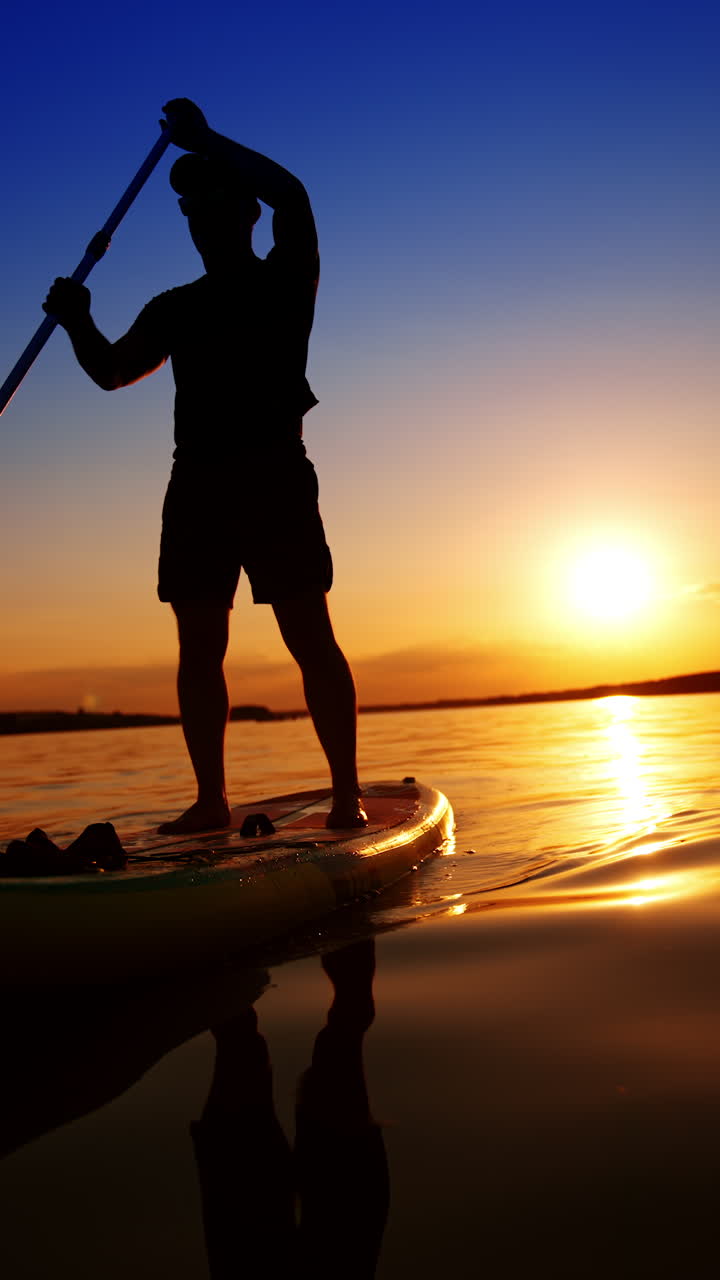 Paddle boarding on the beautiful river at sunset. Man stands on the sup board floating by the water. Low angle view. Vertical video.