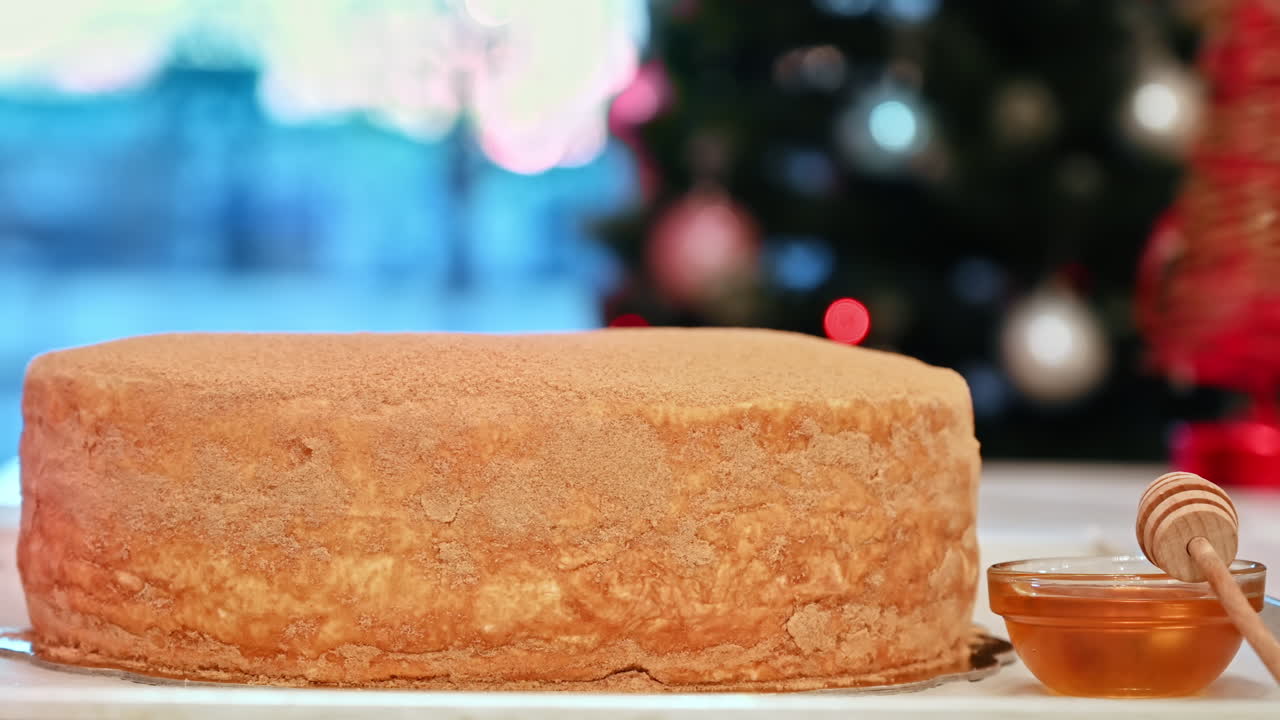 Ready-made cake with a plate of honey near it on a table, Christmas tree on the background