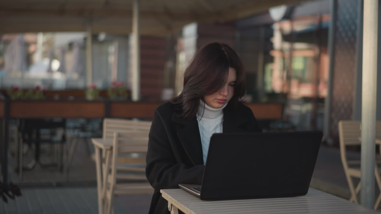 Young woman concentrating on her laptop while working outdoors in urban setting with modern furniture and natural light, background includes blurred urban structures and greenery