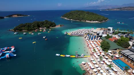 Tourists-Enjoying-White-Sand-Beach,-Sun-Chairs,-Umbrellas,-Emerald-Sea,-Aquapark,-Boats,-and-Affordable-Island-Vacation,-Summer-in-Ksamil