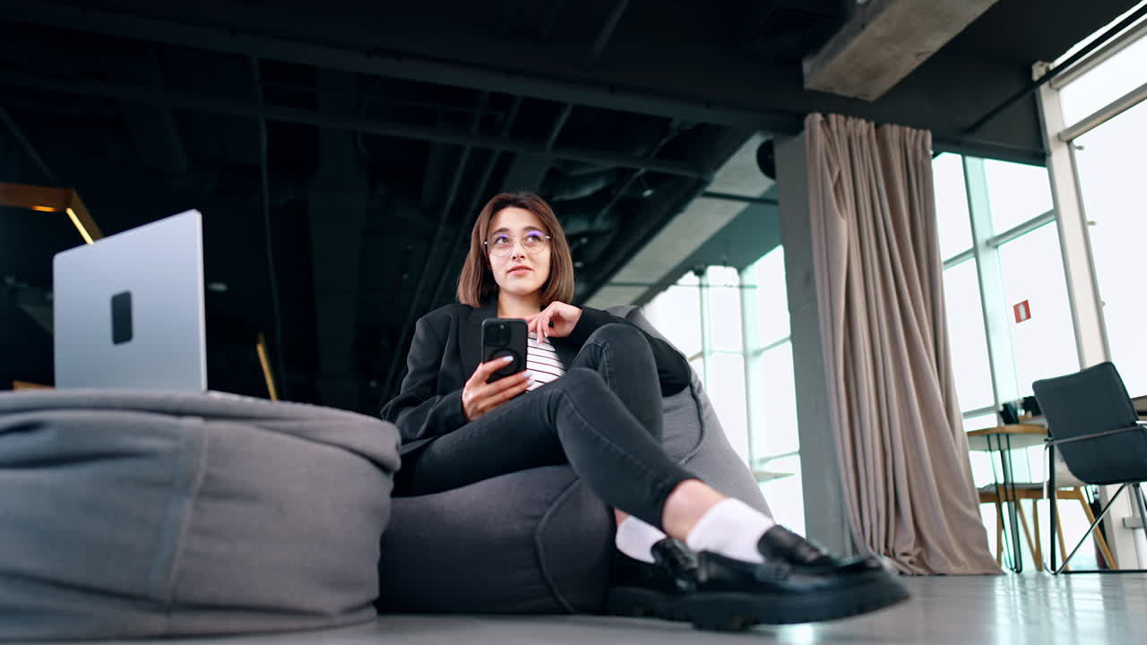 Relaxed brunette woman sits in the bean bag chair holding a phone. Lady looks at the laptop opened in front of her. Low angle view.