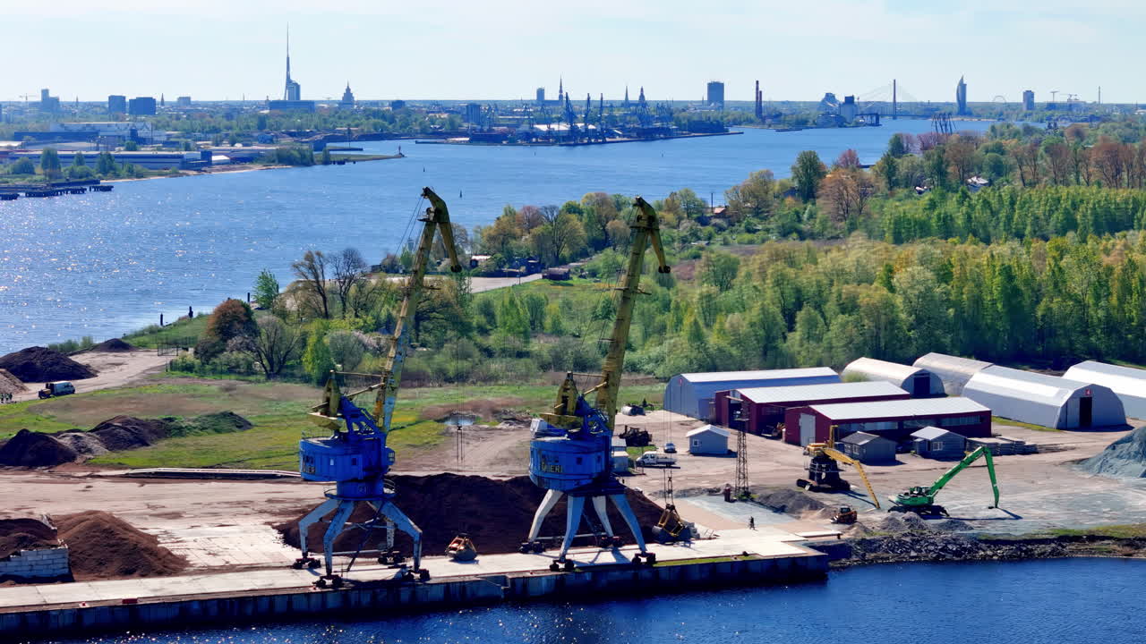 밝고 화창한 늦봄이나 초여름 날, 푸른색과 노란색 갠트리 크레인, 벌크 자재, 창고 건물이 있는 산업 강 항구를 전경으로 포착한 항공 촬영 사진이에요.