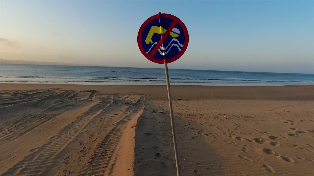 vista en movimiento de un letrero de no nadar en azul amarillo y rojo en una playa de arena desierta con un fondo claro