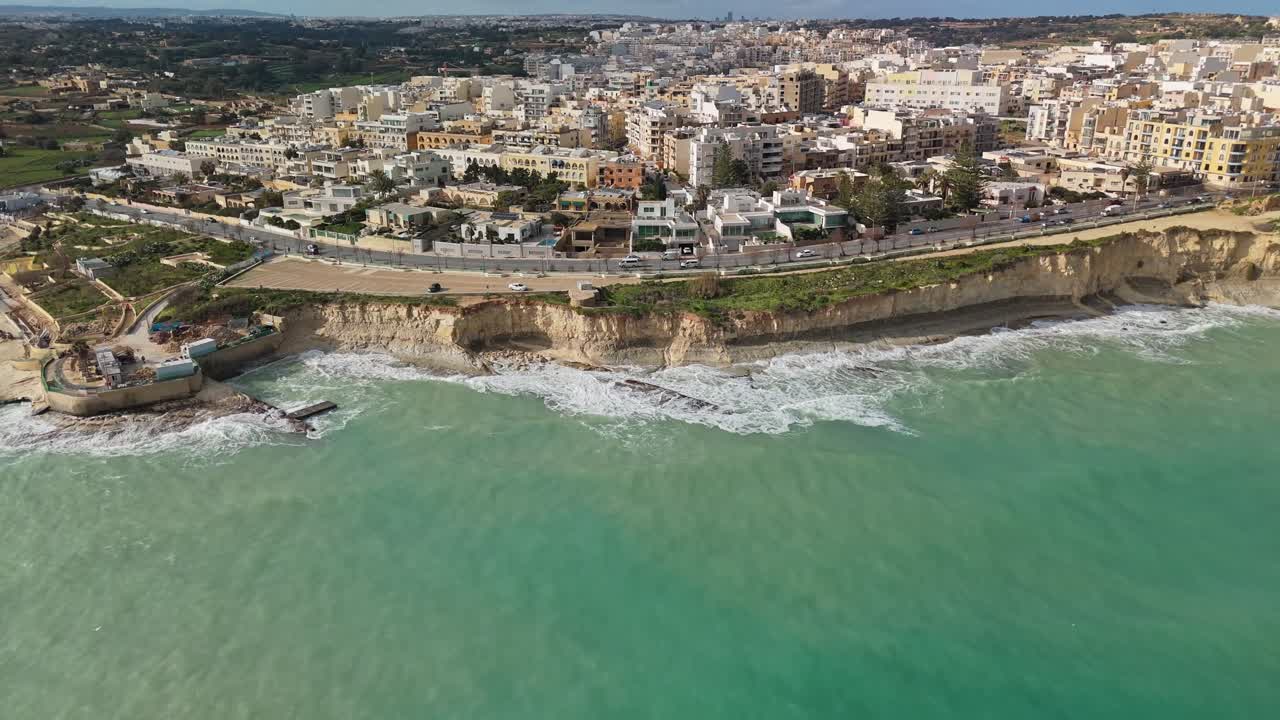 Pemandangan udara mundur yang menanjak di Marsaskala, Malta, menampakkan tebing tepi laut, perairan Mediterania yang bergelombang, arsitektur perkotaan, dan garis pantai musim dingin yang dinamis di siang hari