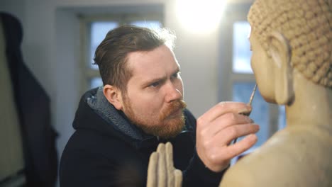 Artista-Masculino-Trabajando-En-La-Cara-De-La-Estatua-De-Buda-Usando-Una-Herramienta-Especial-En-El-Taller.