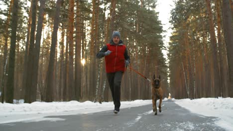 mężczyzna ubrany w zimową odzież sportową biegający z psem po drodze w zaśnieżonym lesie