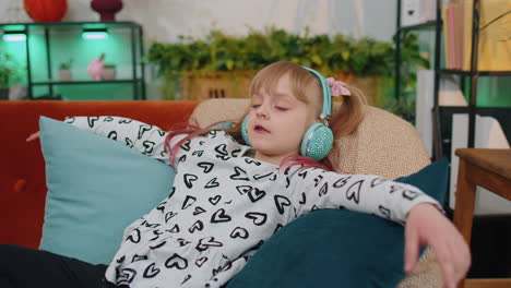 little children girl lying on sofa, wearing wireless headphones, listening to music, singing song