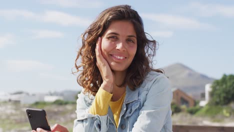 Portrait-of-happy-mixed-race-woman-holding-smartphone-and-smiling-on-sunny-promenade-by-the-sea