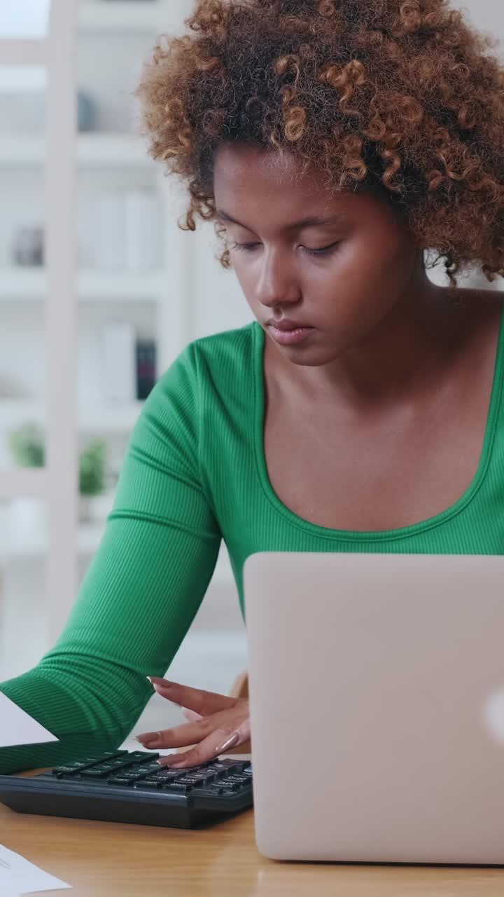 Junge Frau studiert Dokumente, während sie am Schreibtisch zu Hause am Laptop arbeitet
