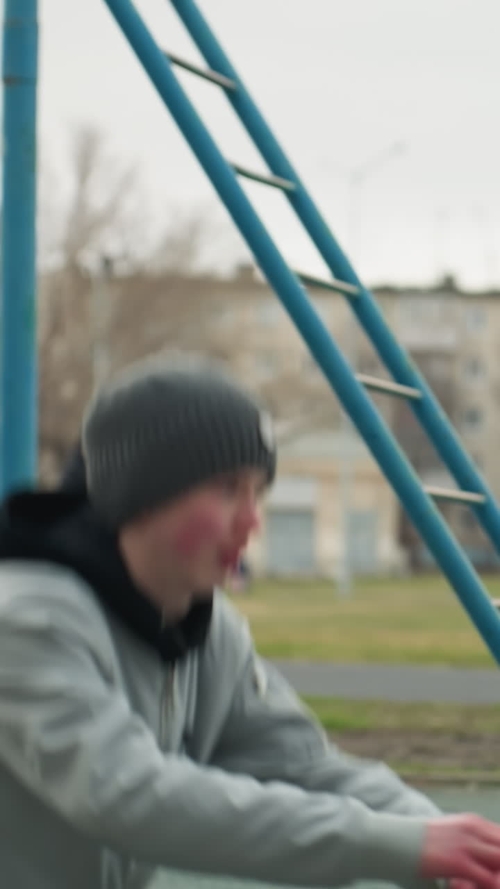 A young boy performs a workout with hands stretched out close to an iron pole, in a stadium with a yellow building in the background, a blurred view of people walking near the building