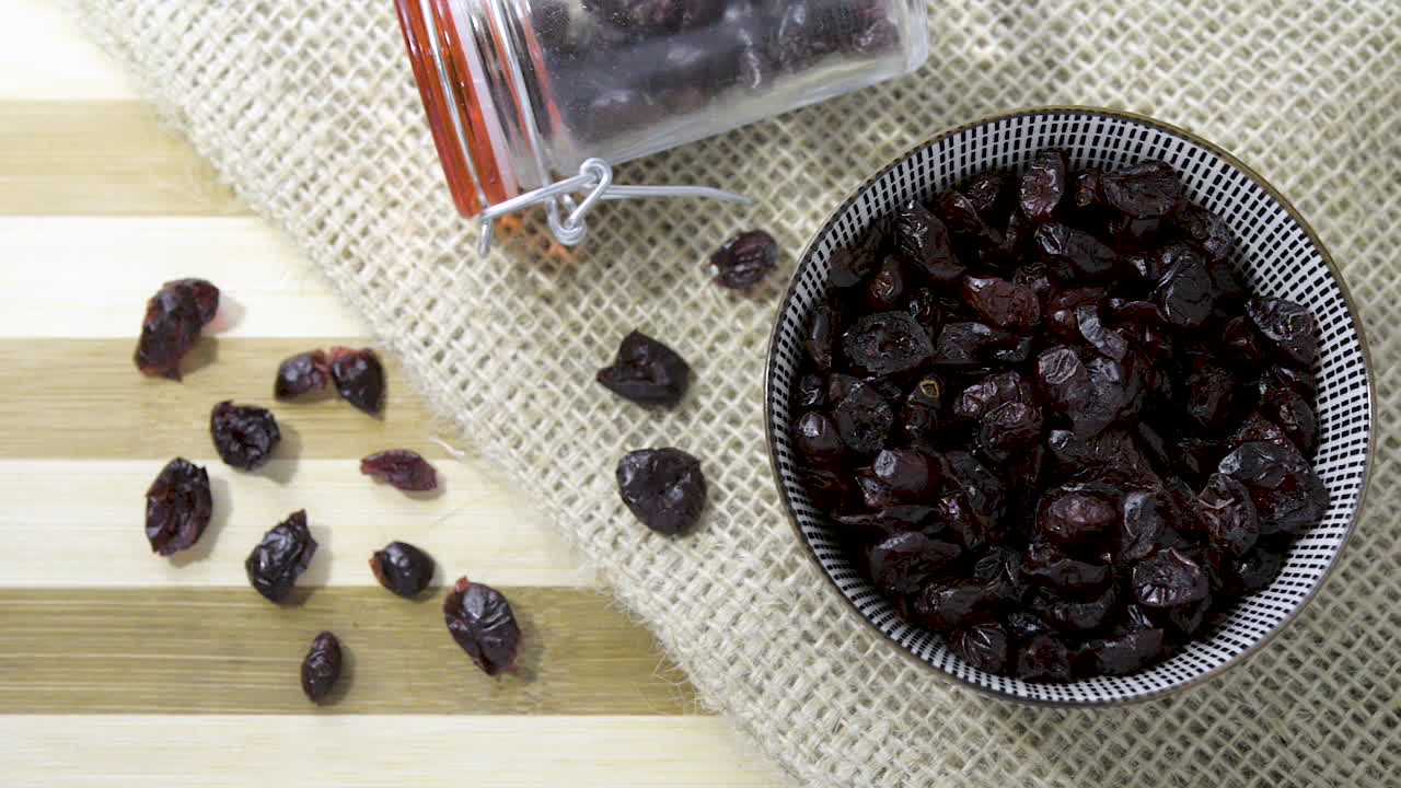 toma panorámica lenta vista de arriba hacia abajo sobre un tazón de cerámica de un frasco de vidrio lleno de una mezcla de castañas con pasas sobre una mesa a rayas con tela de arpillera