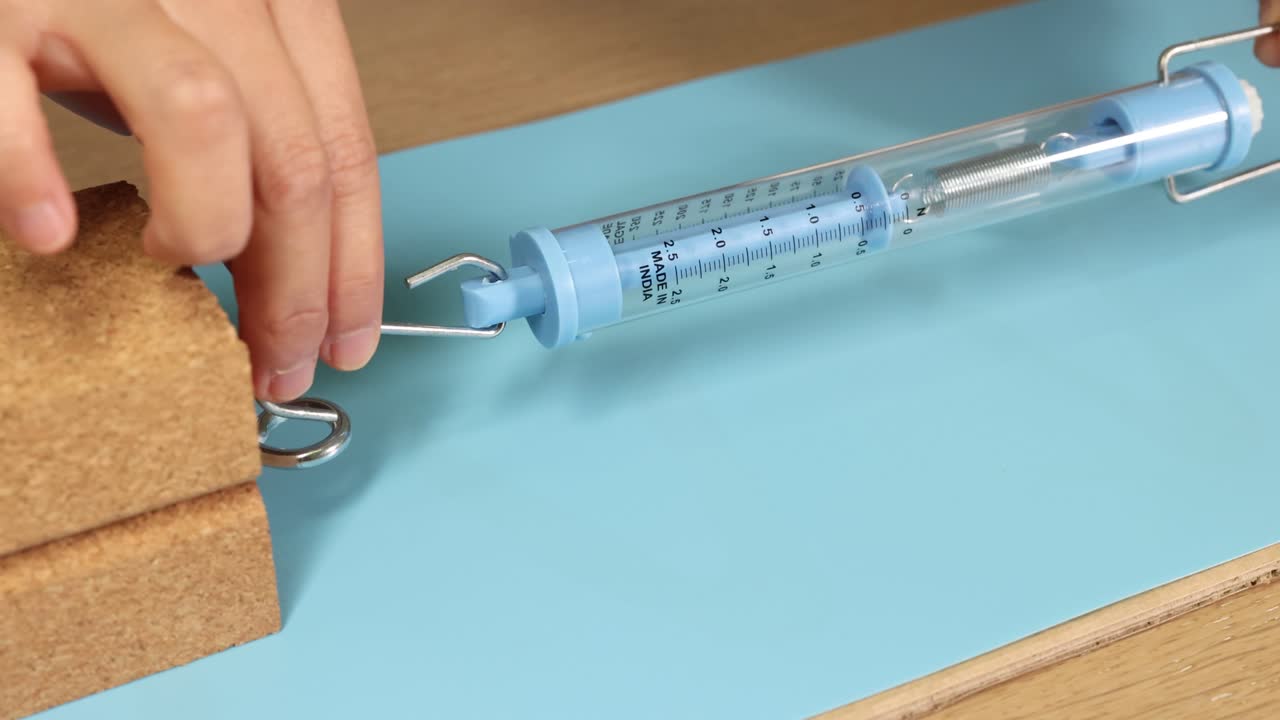 A hand uses a spring scale to measure friction force on a wooden block. Close-up, macro view with clear lighting