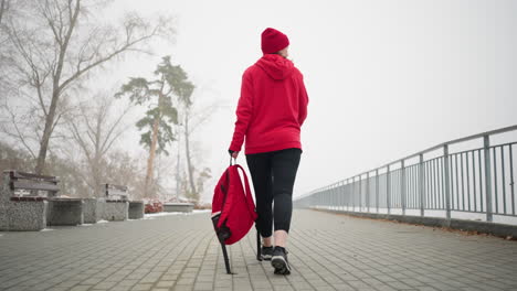 back view of a lady in red winter jacket and head warmer holding a red backpack walking looking away along a fog-covered pathway lined with snowy benches, frosted trees, on a cold winter day