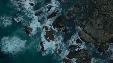 Descending-aerial-top-down-overhead-view-of-big-waves-splashing-on-dark-rocks-on-the-wild-coastline-in-New-Zealand