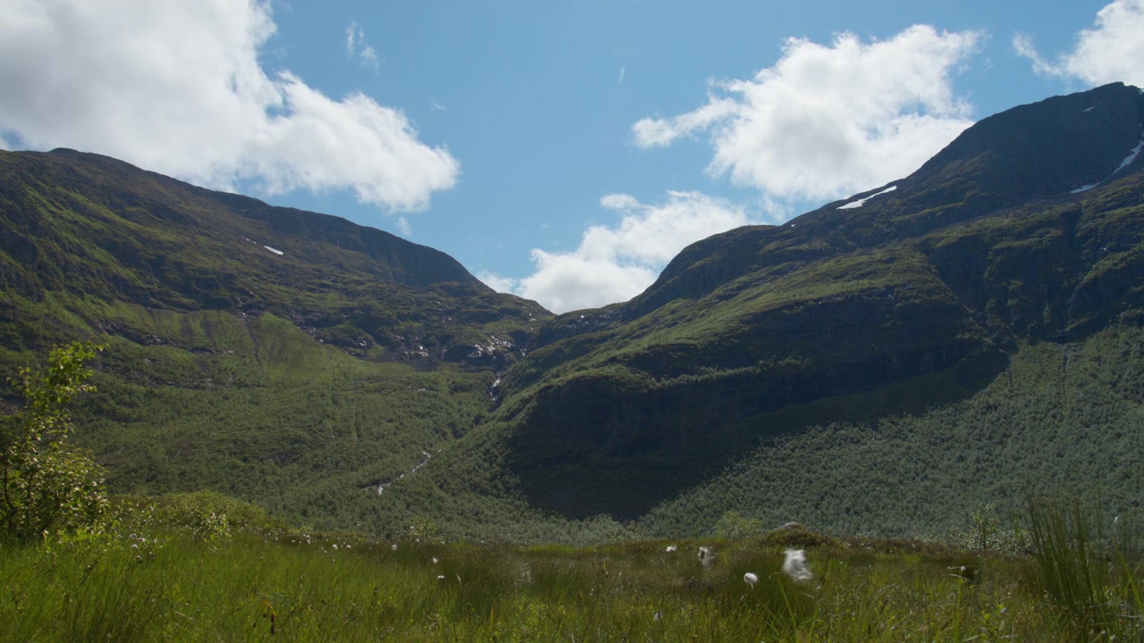 Ampio bacino della valle del fiordo con un primo piano erboso e aspre creste montuose del Sunnmøre sotto un cielo parzialmente nuvoloso.