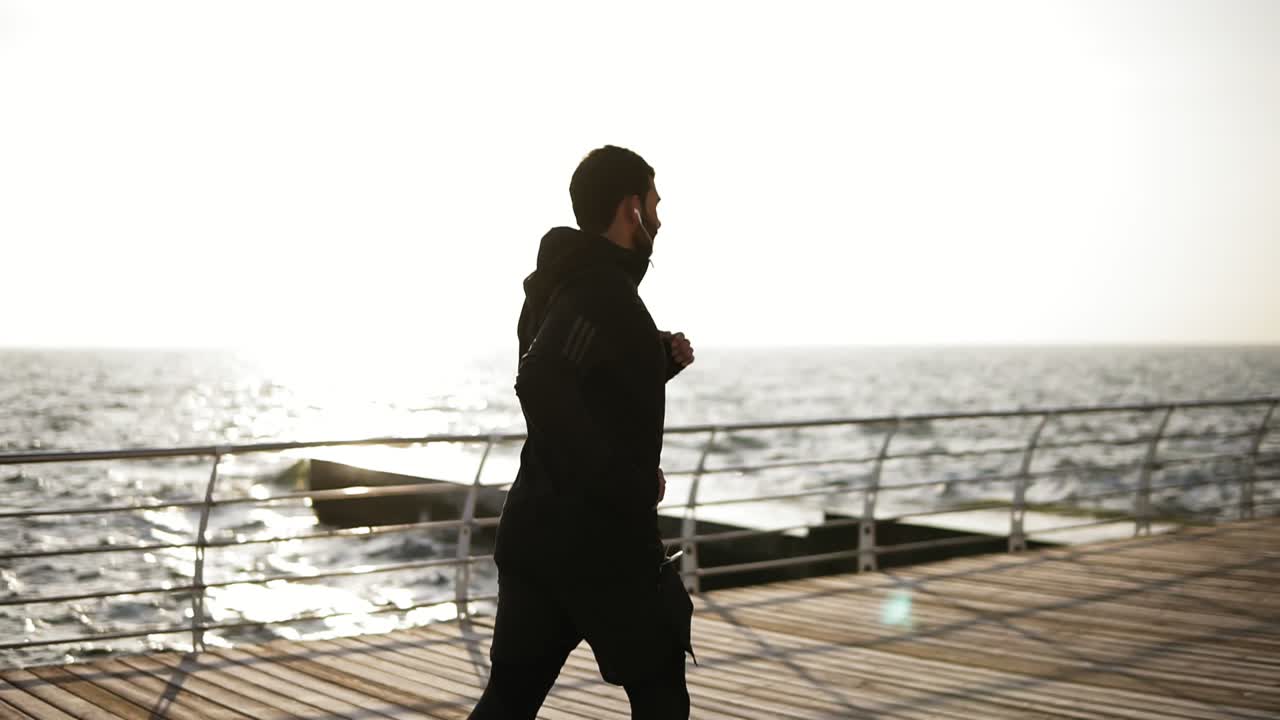 Side view footage of a sportive young male running or jogging on the dock near sea side. Outdoors footage.