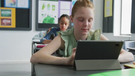 video of caucasian girl sitting at desk using tablet in diverse in school class, copy space
