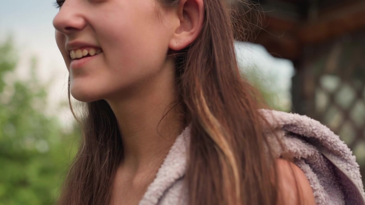 Giovane donna con lunghi capelli castani sistema l'asciugamano all'aperto, passando da un'espressione neutra a un sorriso. La donna indossa abiti chiari in un ambiente esterno con vegetazione e luce soffusa.