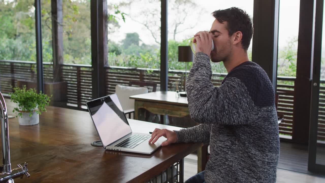 Caucasian man working at home, using laptop and drinking coffee in dining room