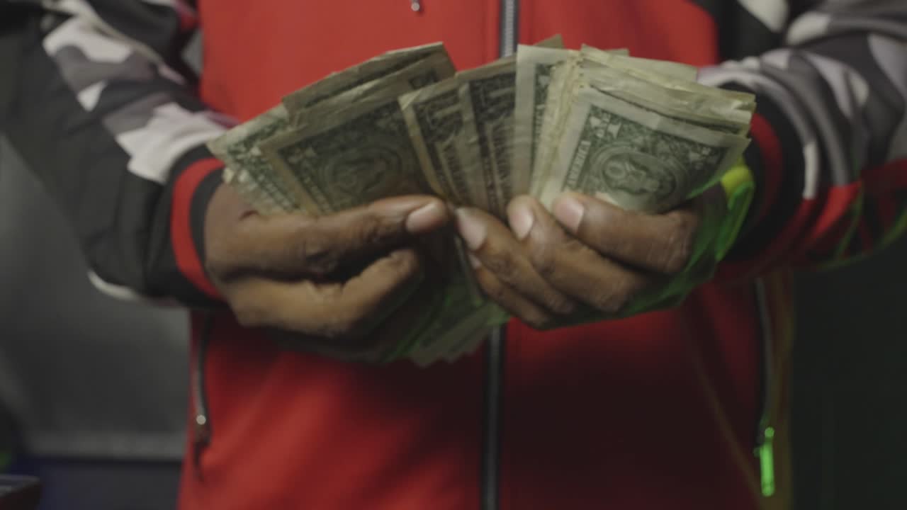 Close-up of hands flipping through stacks of worn dollar bills in a red-and-black jacket, showcasing street hustle and gritty cash flow.