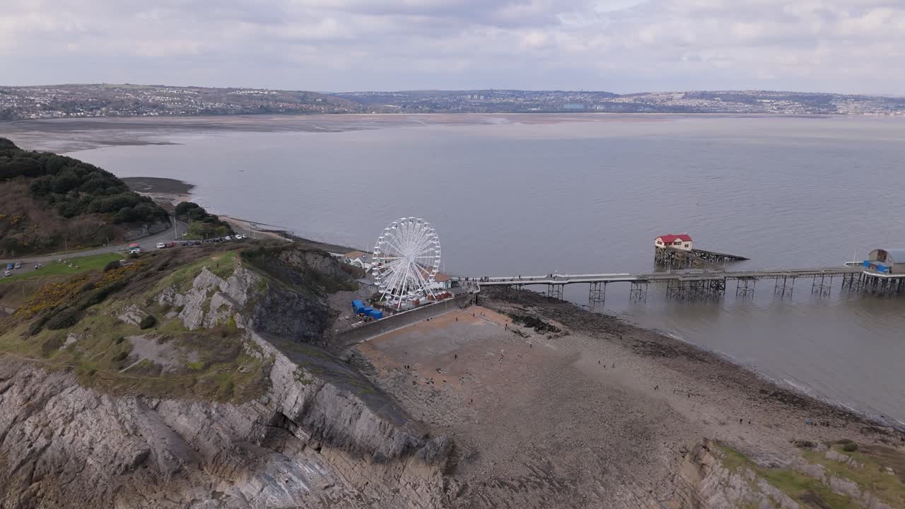 Aerial tracking right above Mumbles Pier in Swansea, UK, with the Big Wheel standing against the cloudy sky