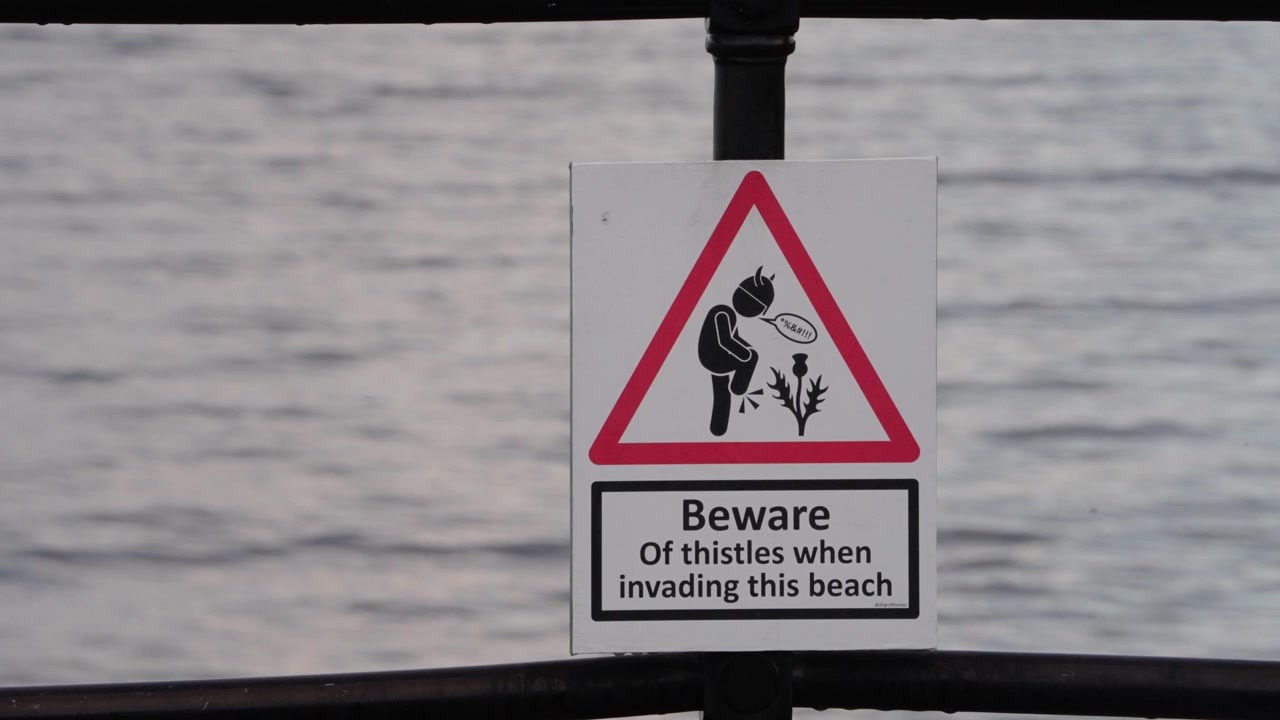 Unique warning signage posted at Largs promenade, Ayrshire, Scotland, cautioning visitors to beware of sharp thistles on the beach, static tripod close-up view