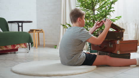 boy sitting on white rug opening old vintage box carefully in cozy living room beside decorated christmas tree, preparing ornaments for holiday decoration in bright peaceful atmosphere