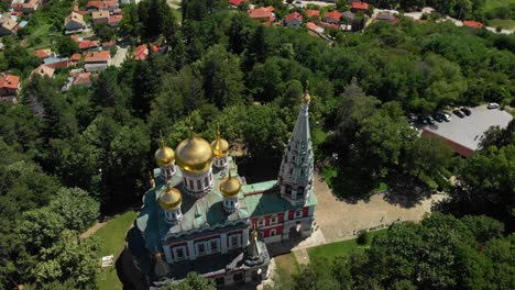 iglesia conmemorativa de shipka en bulgaria - hermosa iglesia de exteriores coloridos y cúpulas doradas con exuberantes árboles verdes circundantes - toma aérea de drones