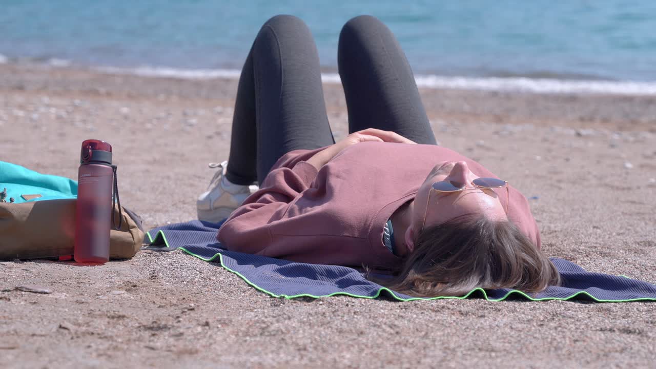 молодая активная женщина греется на солнце и отдыхает после утренней пробежки, лежа на одеяле на песчаном пляже, расслабляясь под звук волн.