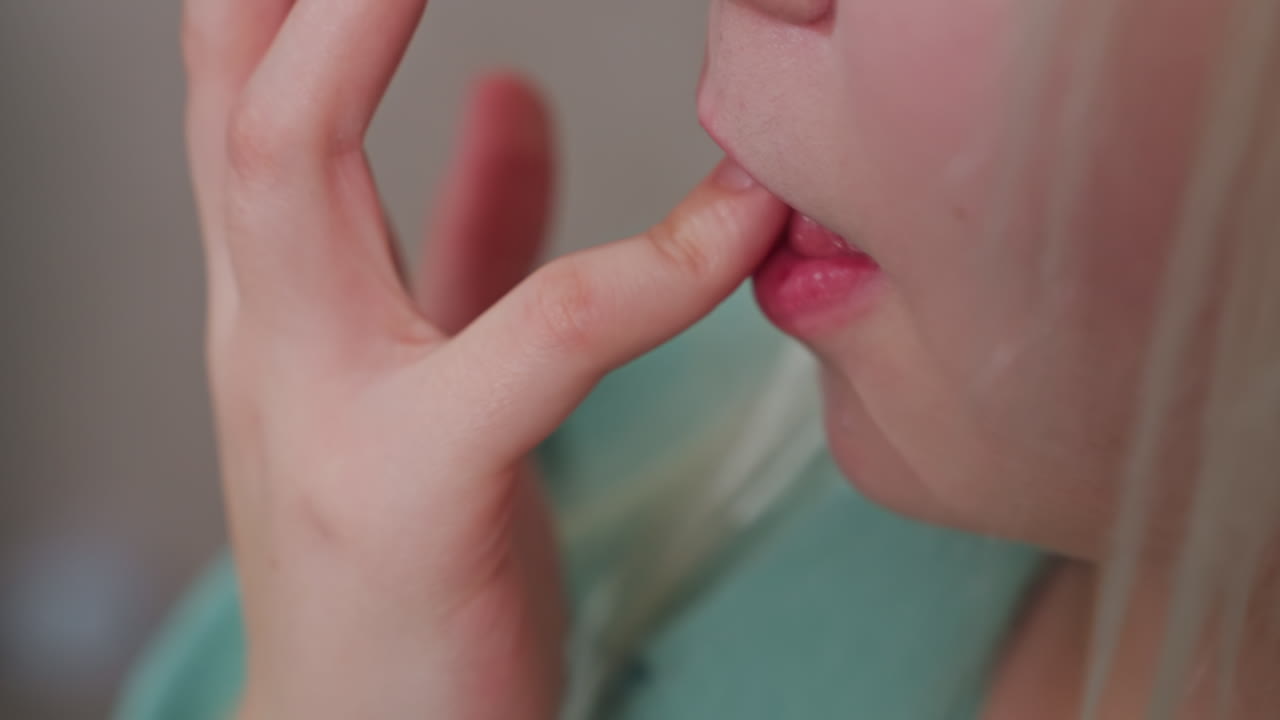 close up of young blonde girl licking finger, natural skin texture and detail visible, soft lighting highlights delicate features, casual and intimate moment indoors with neutral background