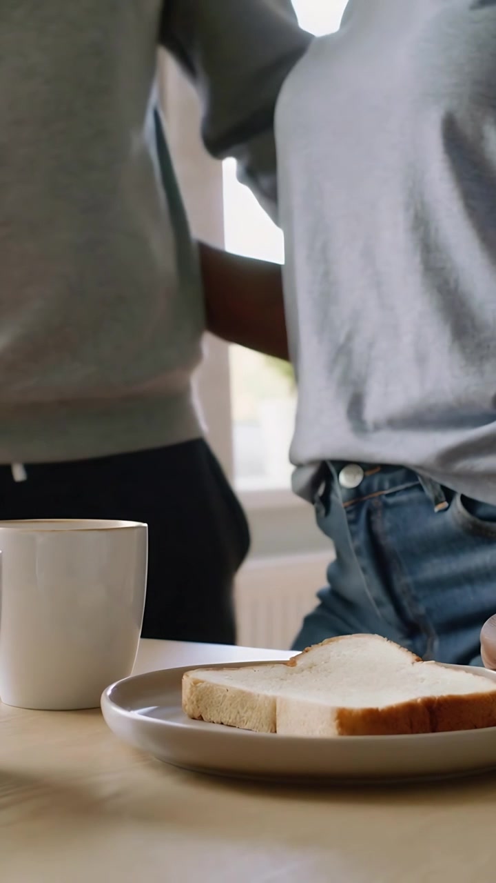 Vertical video: Walking in, two adults in casual wear embracing at kitchen table with mug and bread