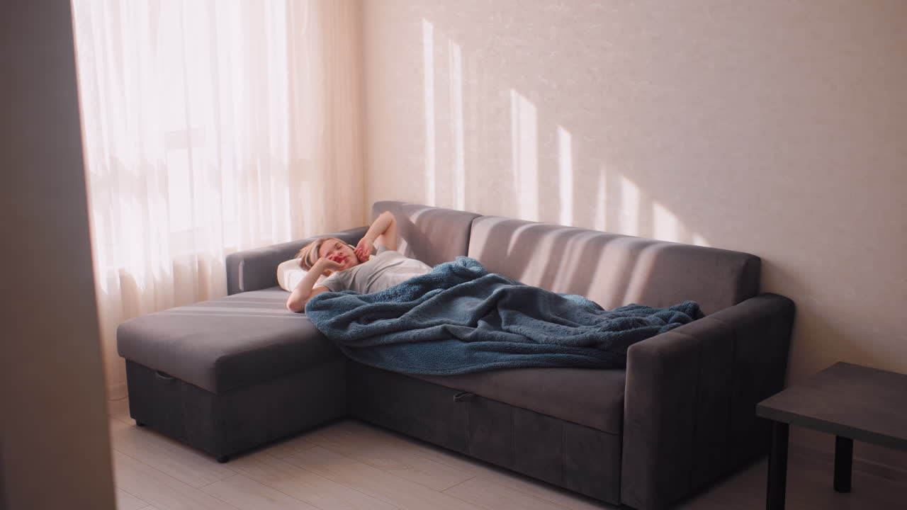 House resident stretches on couch covered with blanket, wiggling leg as morning sunlight streams through curtains, creating cozy peaceful home atmosphere filled with warmth