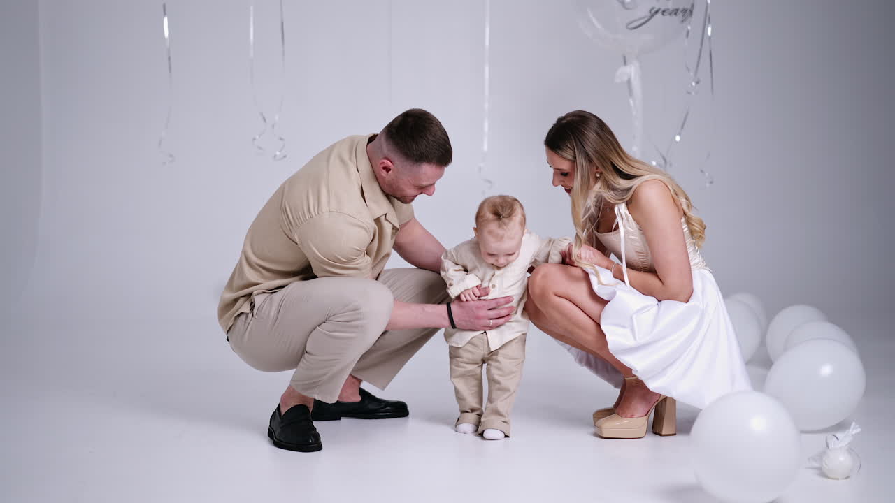 Happy parents hold their baby by the hands walking by the studio. Couple stops and leans to a kid kissing him. Family celebrating first birthday of a child.