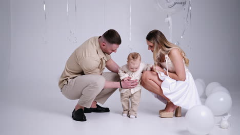 happy parents hold their baby by the hands walking by the studio. couple stops and leans to a kid kissing him. family celebrating first birthday of a child.