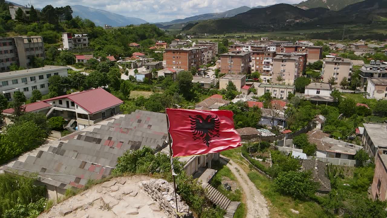toma panorámica de la bandera nacional de albania ondeando en el viento con una pequeña ciudad en el fondo