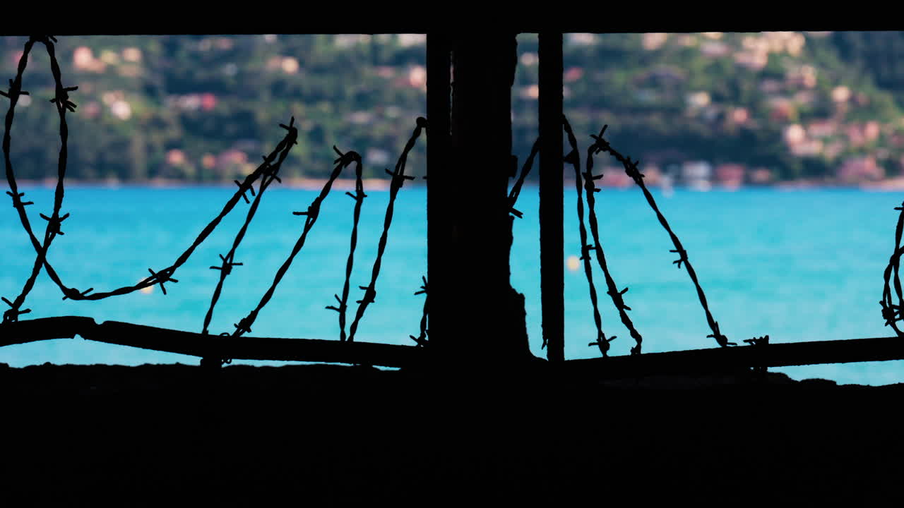 Close up of barbed wire loops along a small window with a blurred view of the sea and a town