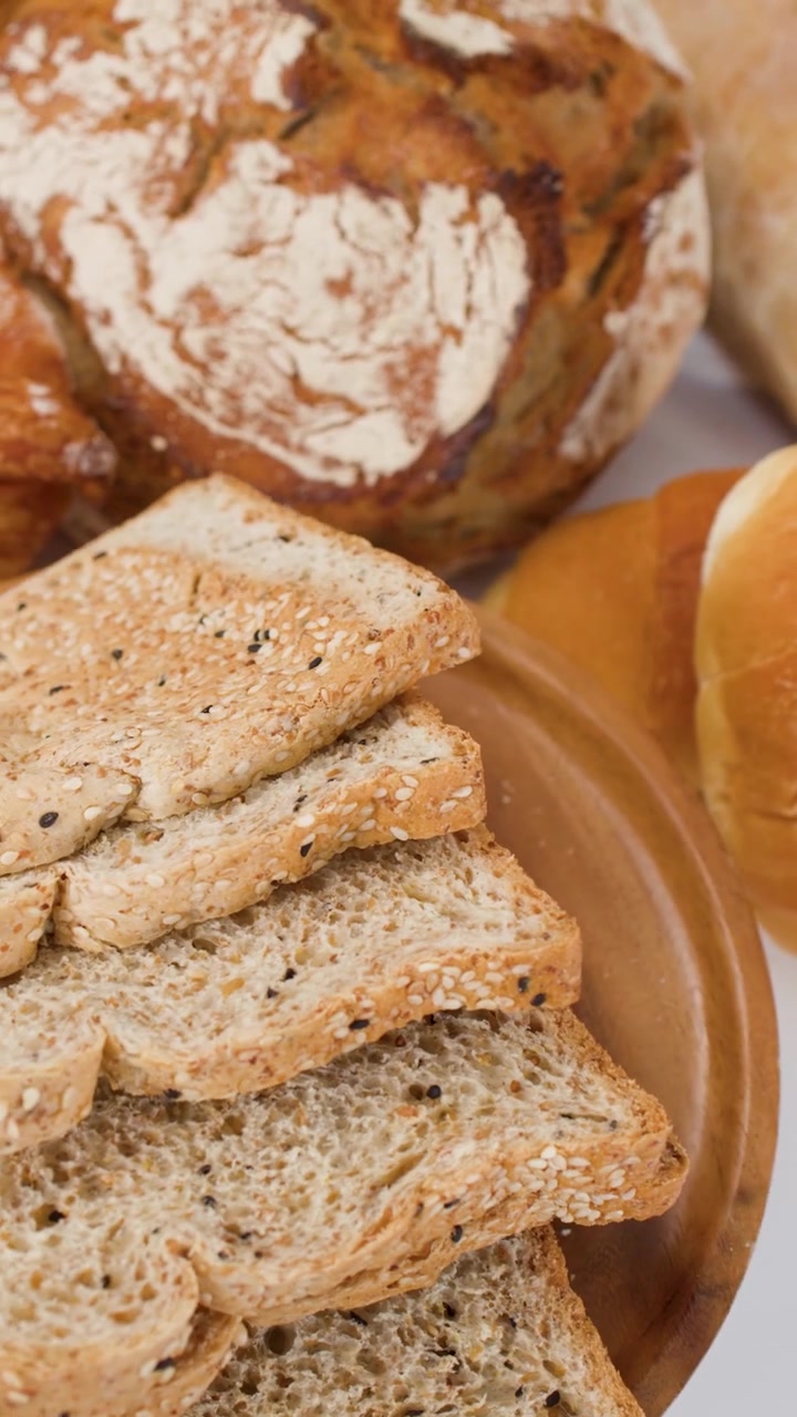 Primo piano rotante di pane affettato ai semi, pane a lievitazione naturale e croissant su un piatto di legno