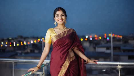 beautiful smiling indian woman wearing traditional sari dress and gold jewelry is posing on a rooftop at dusk with a cityscape in the background