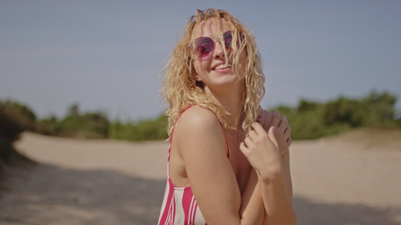Una bella donna bionda con i capelli ricci sorride e ride al rallentatore mentre è in piedi in dune di sabbia bianca e assolate.