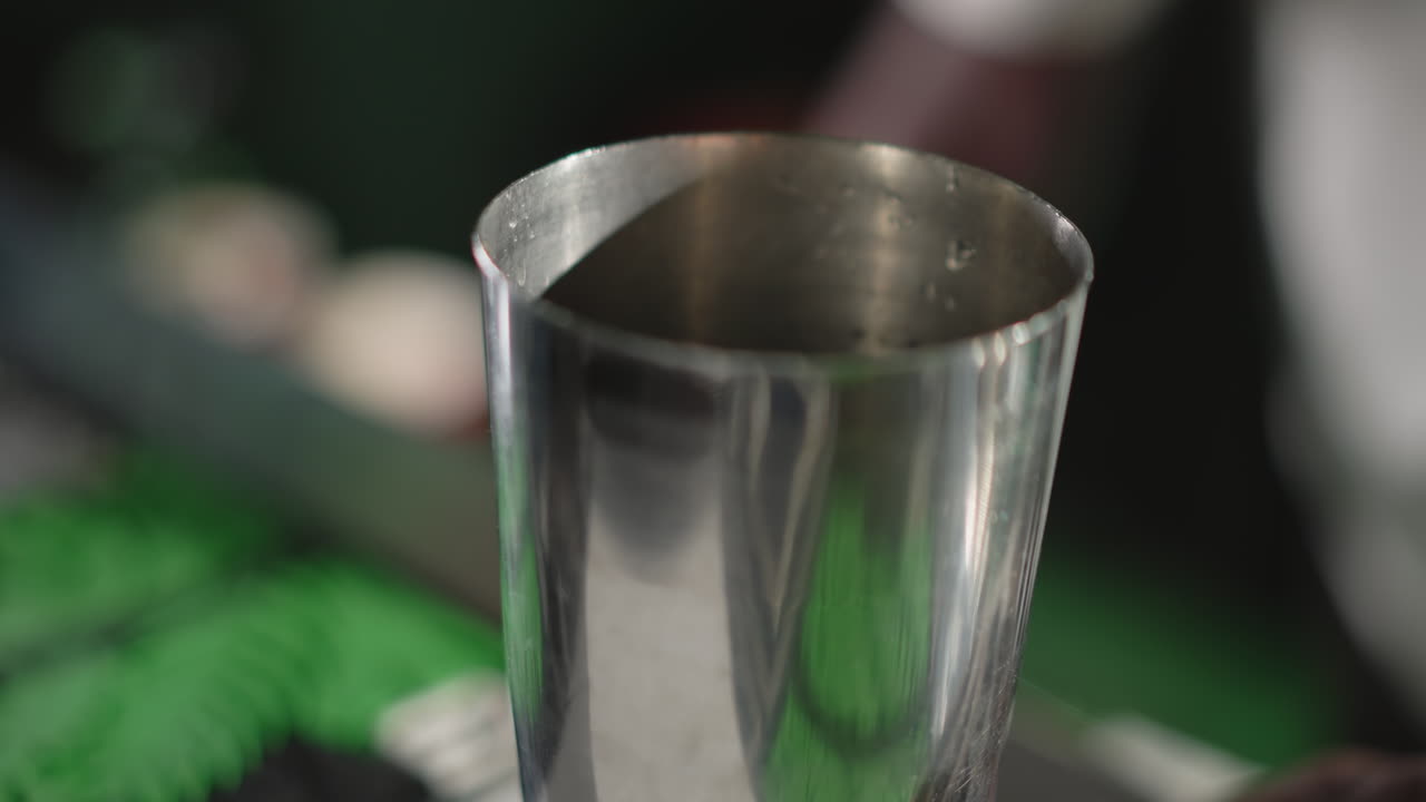 Bartender pouring ice into cocktail shaker, preparing for drink mixing with modern bar tools, focused on drink creation in vibrant green-lit bar setting